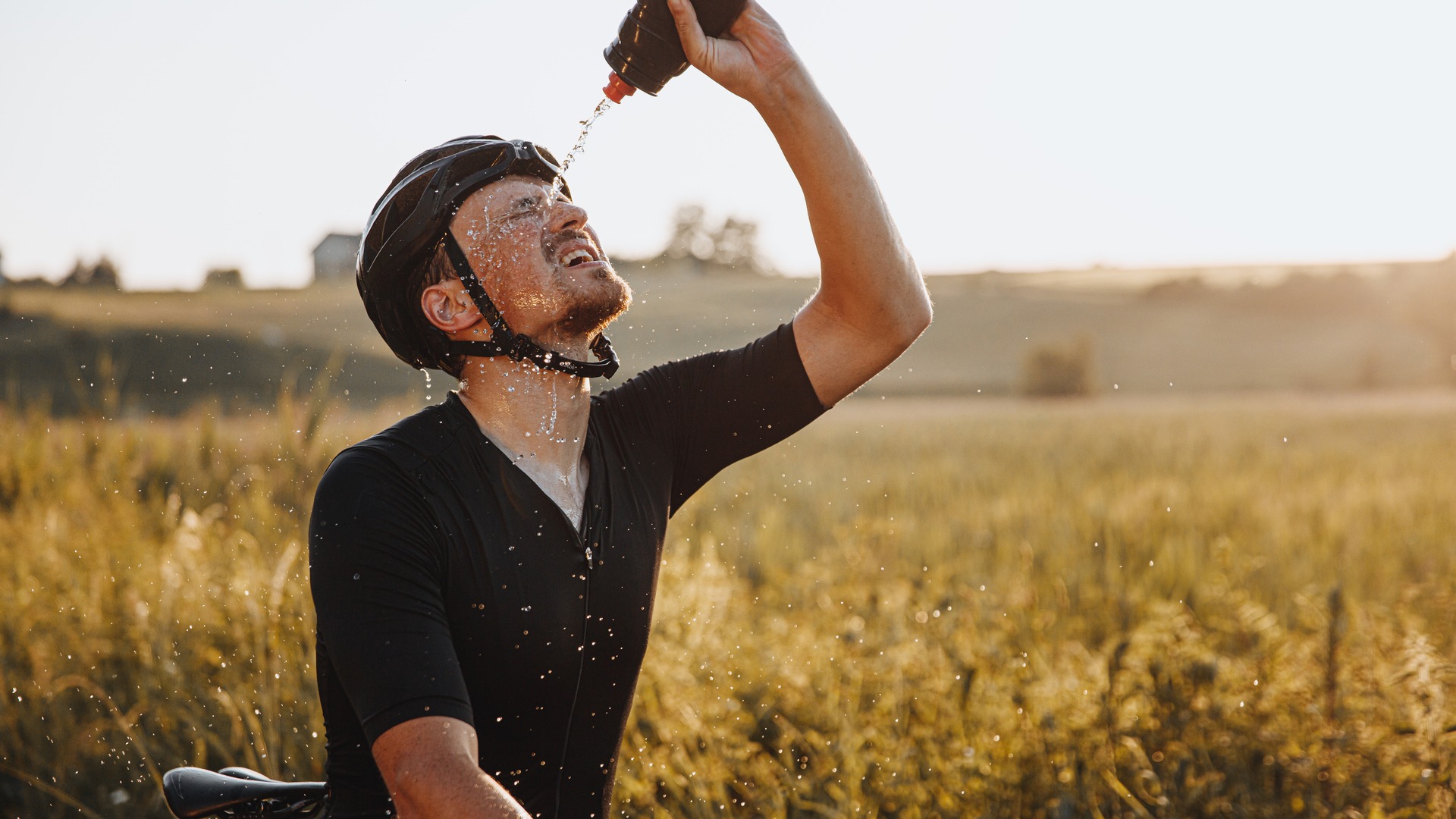 Cyclist pouring water over his face in a sunlit field.