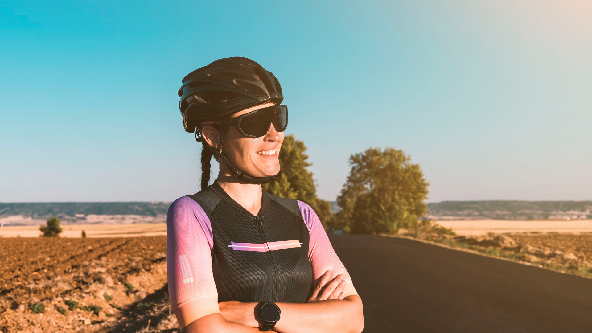 Female cyclist with helmet and sunglasses smiling on an open road with a scenic backdrop, realising the good choice she made to avoid generic cycling training plans
